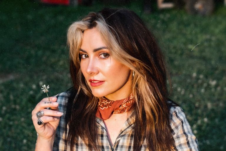 Australian singer Ella Hooper holding a small flower, wearing a neckerchief and plaid shirt outdoors.